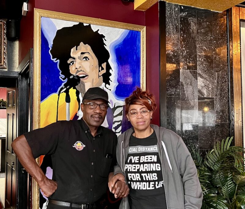 Restaurant owners in dining room with Prince painting behind them