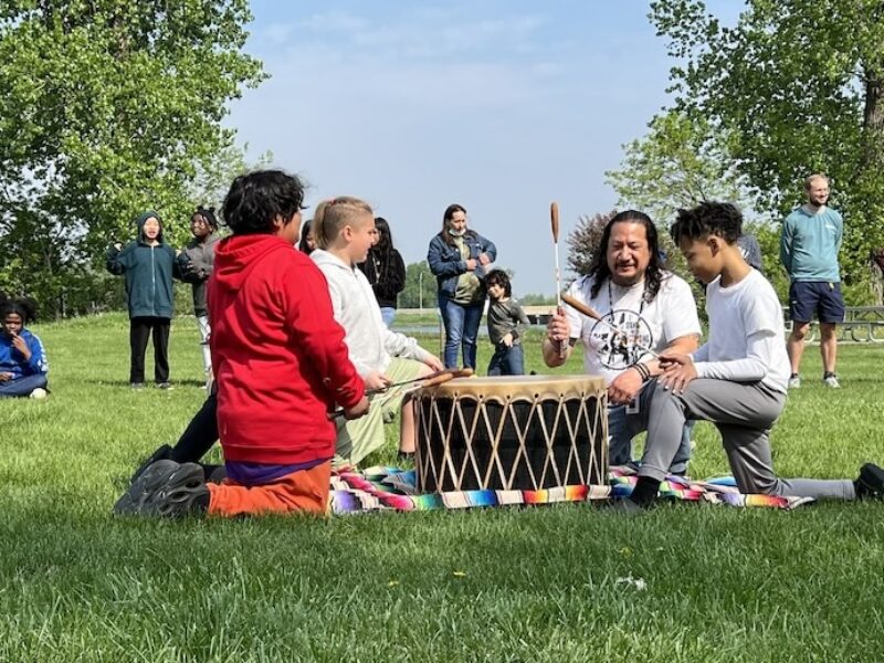 Adult and three kids around a drum playing it for people in the outdoors