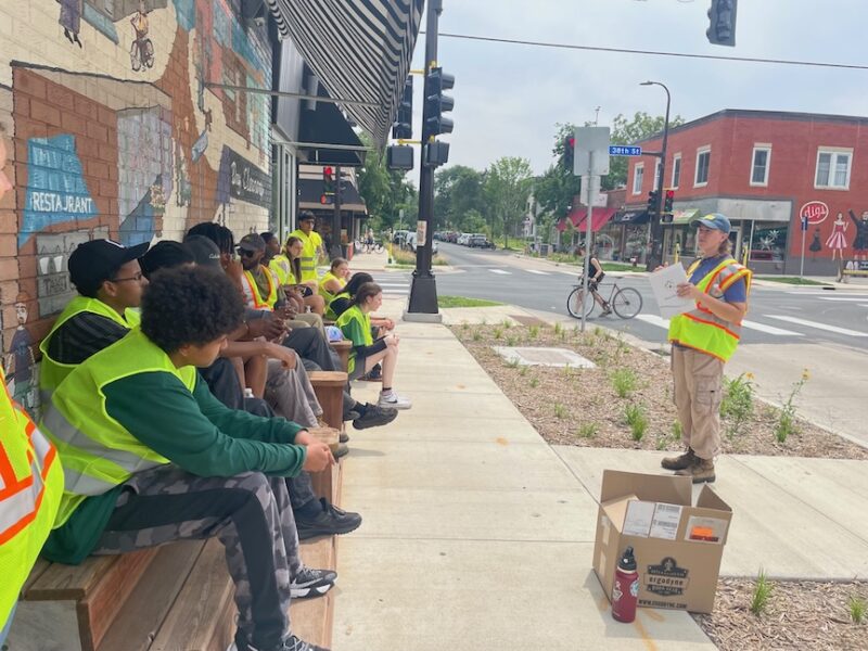 People dressed in attire for outdoor work seated on bench seating in front of storefront listening intently to a speaker